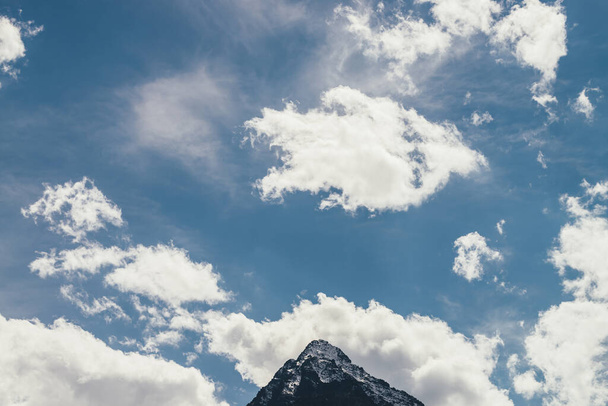 Minimalist cloudscape with pointy peak in cloudy blue sky. Minimal sky landscape with cirrus clouds above peaked top with snow. Beautiful mountain scenery with sharp rock under spindrift clouds. - Photo, Image