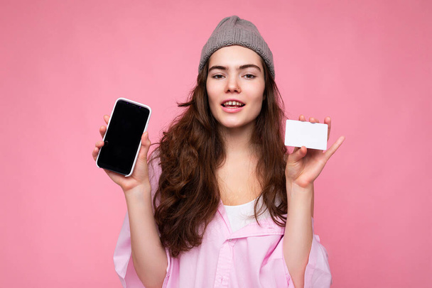 美しい幸せな若い楽しい巻きブルネットの女性はピンクのシャツとグレーの帽子を身に着けていますコピースペースとピンクの背景に隔離され、空の画面表示とクレジットカードで携帯電話を保持 - 写真・画像