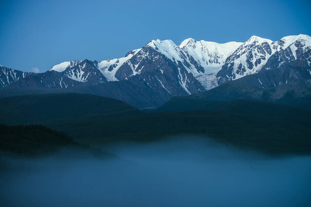Atmosferyczny krajobraz gór z gęstą mgłą i wielkim szczytem górskim śniegu pod półmrokiem nieba. Alpejska sceneria z dużymi śnieżnymi górami nad gęstą mgłą w nocy. Szczyt śniegu nad chmurami o zmierzchu. - Zdjęcie, obraz