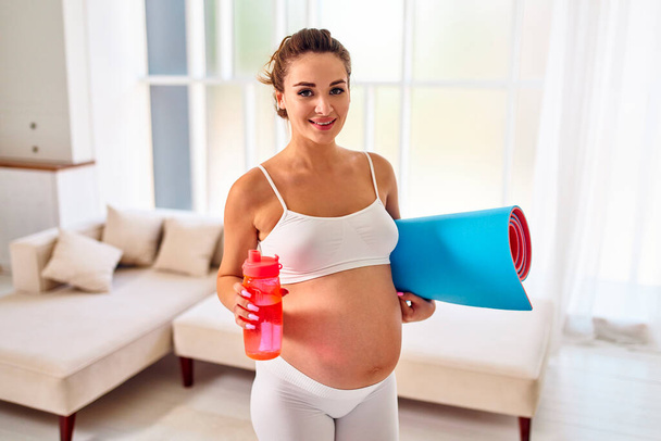 La joven embarazada en ropa deportiva sostiene una alfombra de goma y bebe agua de una botella en la sala de estar. Deportes y un estilo de vida saludable durante el embarazo. - Foto, Imagen