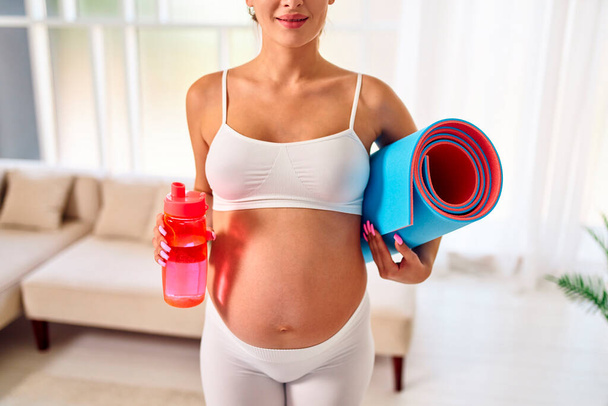 Junge schwangere Frau in Sportbekleidung hält eine Gummimatte in der Hand und trinkt Wasser aus einer Flasche im Wohnzimmer. Sport und ein gesunder Lebensstil während der Schwangerschaft. - Foto, Bild