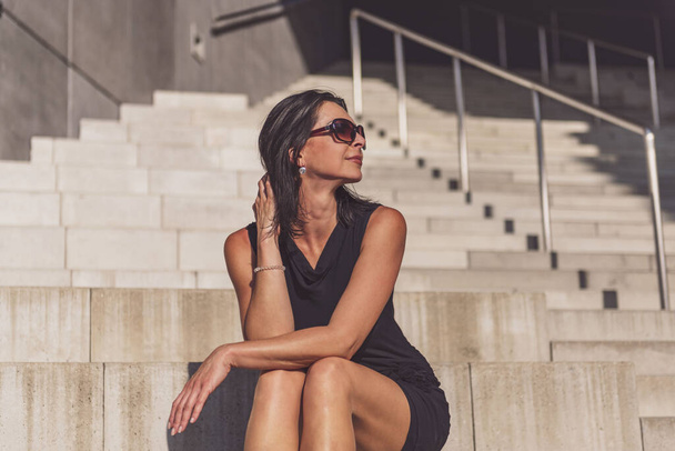 Dark-haired woman posing in a black dress on the concrete stairs. The woman is wearing black sunglasses. It's a beautiful sunny day. - Fotó, kép