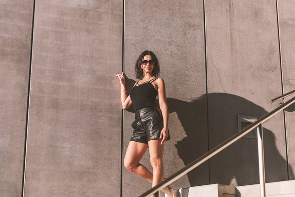 Mulher de cabelos escuros posando em um vestido preto nas escadas de concreto. A mulher está a usar óculos escuros. Está um belo dia de sol.. - Foto, Imagem
