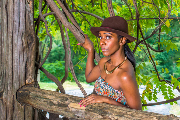 В коричневой шляпе, с золотым кольцом на шее, красивая черная женщина стоит снаружи в маленьком лесу и смотрит на тебя - Фото, изображение