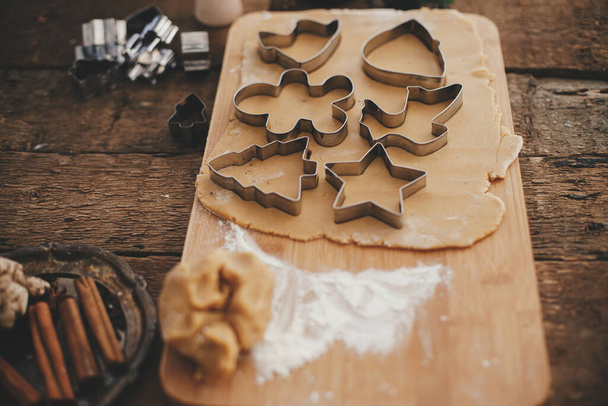 Ciasto piernikowe z bożonarodzeniowymi metalowymi frezami na drewnianej desce. Atmosferyczny obraz nastrojów. Dokonywanie tradycyjnych świątecznych pierników ciasteczka na rustykalnym stole z przyprawami i dekoracjami - Zdjęcie, obraz