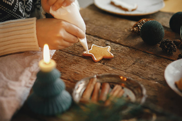 Dłonie dekorowanie Christmas piernik cookie gwiazda z lukrem na rustykalnym stole ze świecą, przyprawy, dekoracje. Atmosferyczny obraz. Robiąc tradycyjne pierniki świąteczne ciasteczka - Zdjęcie, obraz
