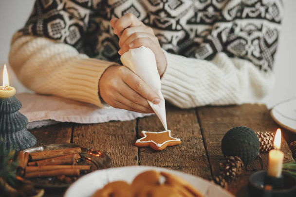 Χέρια διακόσμηση χριστουγεννιάτικο μπισκότο αστέρι μπισκότο με γλάσο σε ρουστίκ τραπέζι με κερί, μπαχαρικά, διακοσμήσεις. Ατμοσφαιρική κυκλοθυμική εικόνα. Γυναίκα κάνει παραδοσιακά μπισκότα χριστουγεννιάτικο μελόψωμο - Φωτογραφία, εικόνα