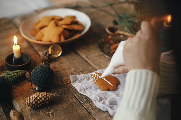Hände dekorieren Lebkuchen-Weihnachtsbaum mit Zuckerguss auf rustikalem Tisch mit Serviette, Kerze, Gewürzen, Dekoration. Stimmungsvolles Stimmungsbild. Lebkuchen backen - Foto, Bild