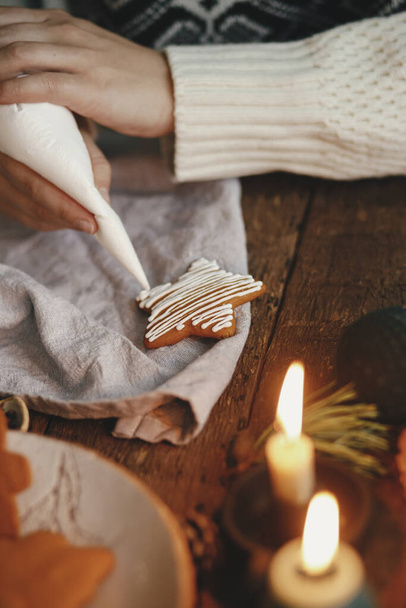 Hände dekorieren weihnachtlichen Lebkuchenstern mit Zuckerguss auf rustikalem Tisch mit Kerzen, Gewürzen, Dekorationen. Stimmungsvolles Stimmungsbild. Lebkuchen backen - Foto, Bild