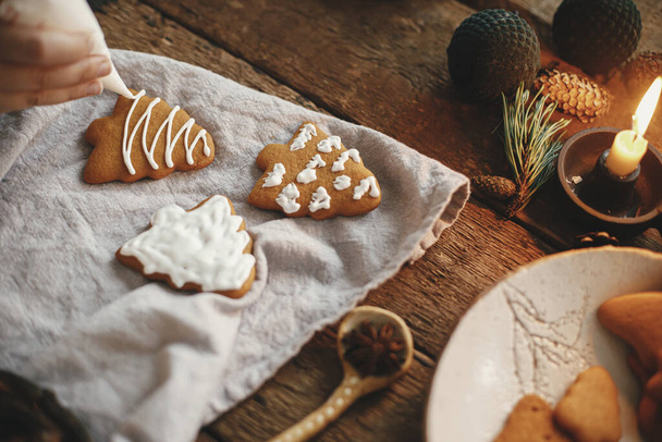 Mãos decorando biscoito de gengibre árvore de Natal com cobertura na mesa rústica com guardanapo, vela, especiarias, decorações. Imagem temperamental atmosférica. Fazendo biscoitos tradicionais de gengibre de Natal - Foto, Imagem