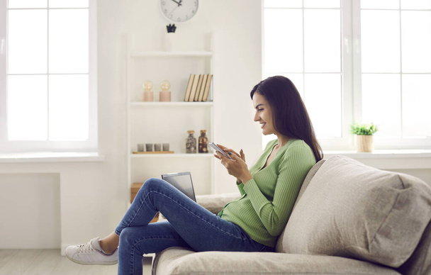 Happy young casual woman relaxing at home in living room sitting on sofa with mobile phone. - Foto, immagini