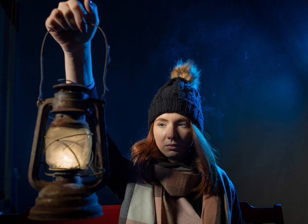 Pretty red hair girl with blue eyes, wearing cap, sitting on bench and holding lantern in dark  - Foto, afbeelding