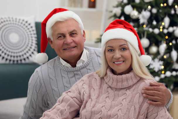 Happy mature couple in Santa hats at home. Christmas celebration - Фото, изображение