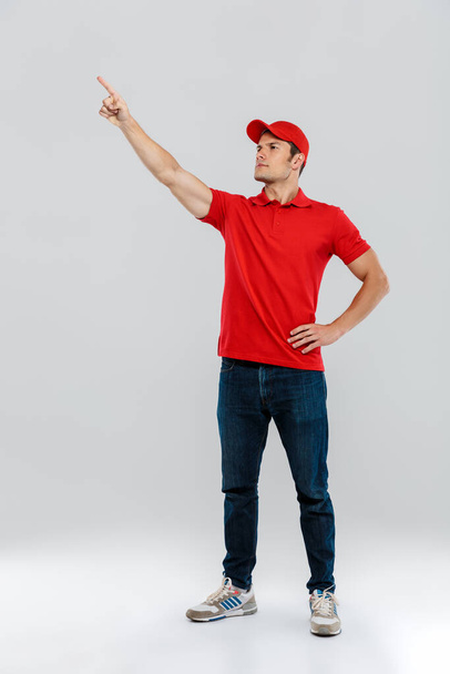 Young delivery man wearing uniform looking and pointing finger aside isolated over white wall - Photo, Image