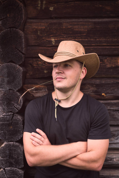 Young man smiling in a cowboy hat on the background of a wooden log house. - Фото, зображення