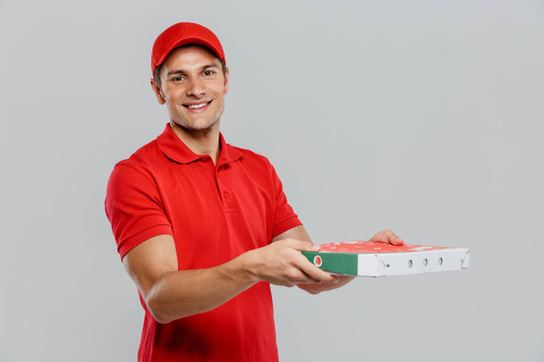 Young delivery man in hat smiling while posing with pizza box isolated over white wall - Foto, imagen