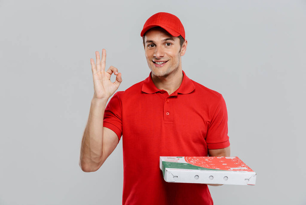 Young delivery man in hat gesturing while posing with pizza box isolated over white wall - Фото, изображение