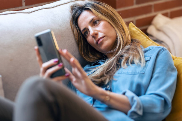Shot of pretty mature woman using mobile phone while relaxing sitting on a couch at home - Photo, Image