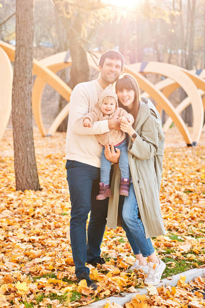 かわいい1歳の女の子と彼女の若い両親の肖像画。黄色の秋の公園を歩く。葉は秋と黄色の葉。晴れた日。お父さんとお母さん. - 写真・画像