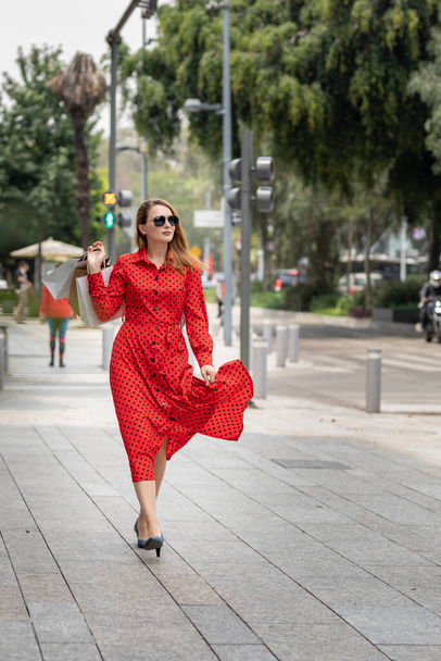 Young happy Caucasian woman in red dress walking stylishly down the street carrying shopping bags - Foto, imagen