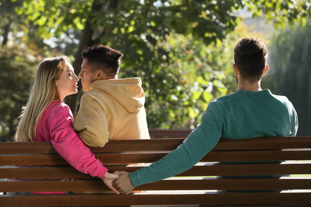 Woman holding hands with another man behind her boyfriend's back on bench in park. Love triangle - Photo, image