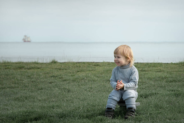 Small blond child sits on grass and smiles on the seashore - Fotoğraf, Görsel