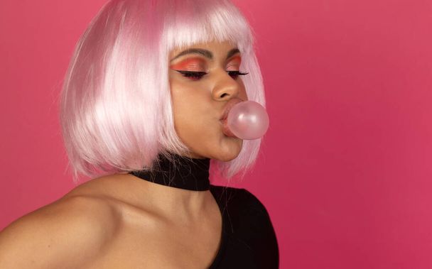 sexy joven afroamericana mujer con vívido maquillaje inflando una burbuja de goma de mascar sobre un fondo rosa - Foto, imagen
