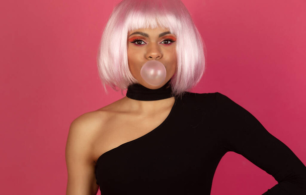 sexy young African American woman with vivid make up inflating a bubble of gum on a pink background - Fotó, kép