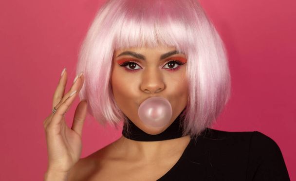 sexy young African American woman with vivid make up inflating a bubble of gum on a pink background - Foto, imagen
