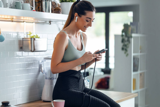 Shot of sporty woman listening to music with her smartphone whiledrinking coffee in the kitchen at home. - Fotó, kép