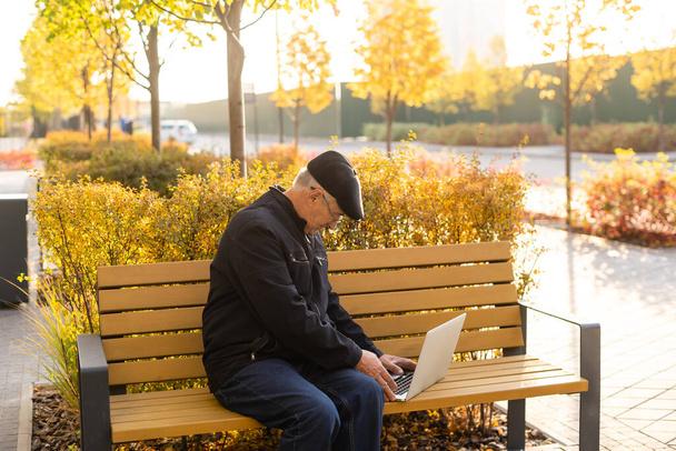 Starszy mężczyzna używa laptopa w parku. Przystojny starszy pan w okularach pracuje na zewnątrz. - Zdjęcie, obraz