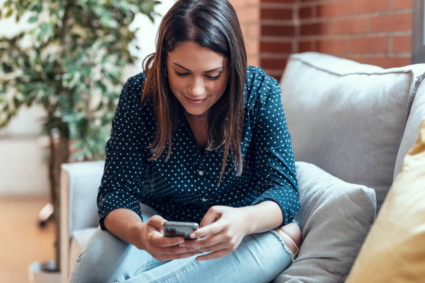 Tiro de mulher bonita enviando mensagens com seu telefone celular enquanto sentado em um sofá em casa. - Foto, Imagem