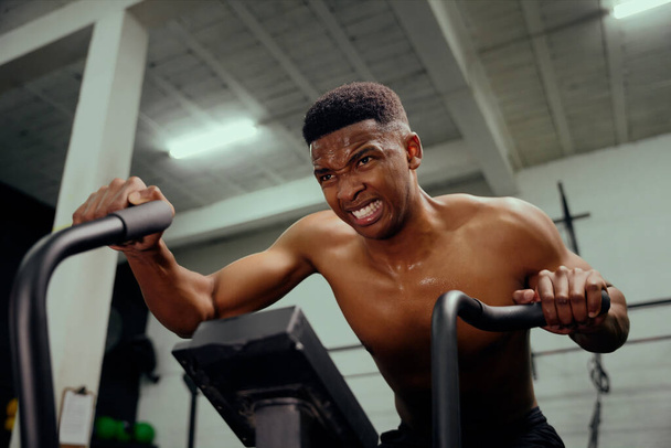 African American male using an elliptical trainer doing intense training. Male athlete exercising intensely in the gym. High quality photo - Photo, Image