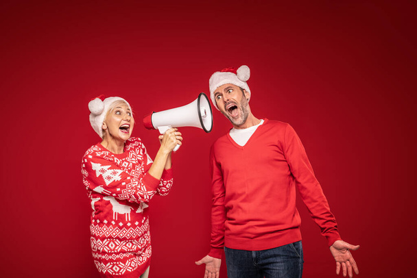 Couple d'âge mûr en chandails de Noël et chapeaux santa claus criant sur mégaphone. Fond rouge du studio. Beaucoup d'espace pour copier. Concept de Noël - Photo, image