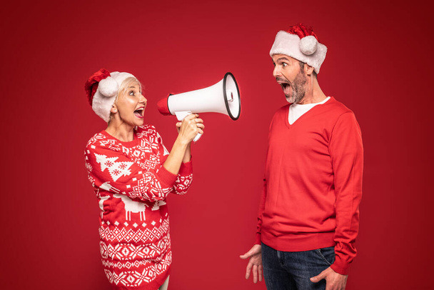 Mature couple in christmas sweaters and santa claus hats shouting on megaphone. Red studio background. A lot of copy space. Xmas concept - Photo, image
