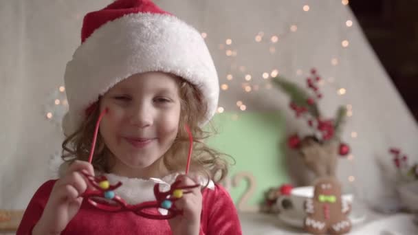 Wazig klein meisje met kerstmuts zet een kerstbril op, kijkt naar de camera en glimlacht. Xmas presenteert vakantie kindertijd concept. Genieten van de kersttijd - Video