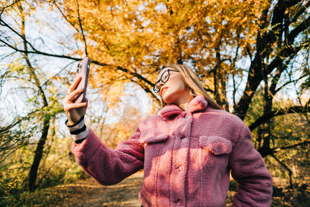 Young woman making self photo and enjoying autumn weather in the park. Woman walking in the autumn park. - Fotoğraf, Görsel