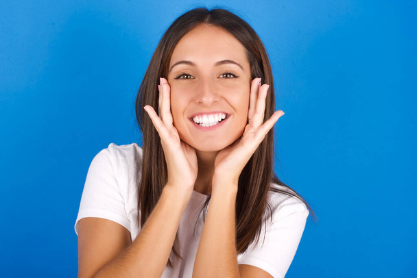 Счастливая красивая молодая женщина нежно касается обеих щек, нежно улыбается, показывает белые зубы, позитивно смотрит в камеру, - Фото, изображение