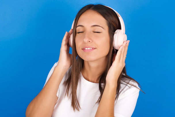 beautiful young woman with headphones on her head, listens to music, enjoying favourite song with closed eyes, holding hands on headset. - Photo, image