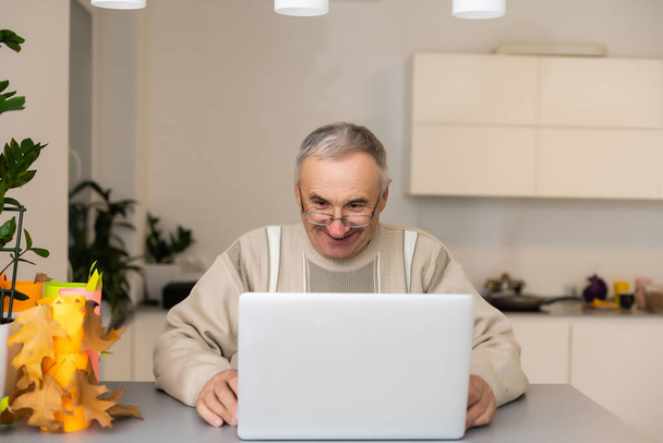 Przystojny starzec ubrany w luźnym stylu formalnym za pomocą laptopa, siedząc na kanapie w domu. Szczęśliwy dziadek komunikuje się z wnukami. Koncepcja połączenia wideo. - Zdjęcie, obraz