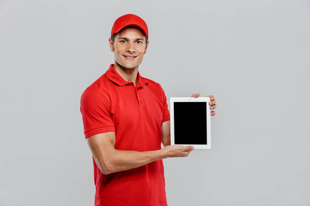 Young delivery man in uniform smiling while showing tablet computer isolated over white wall - Foto, Imagem