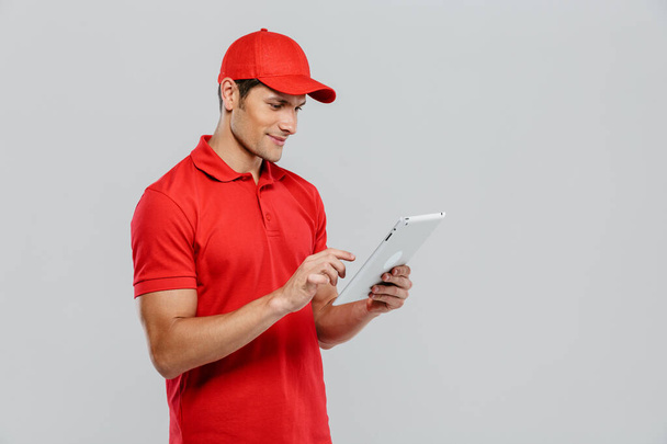 Young delivery man in uniform smiling while using tablet computer isolated over white wall - Фото, зображення
