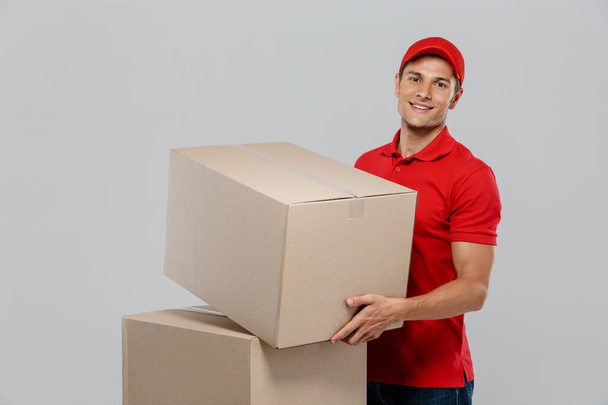 Young delivery man in hat smiling while posing with cardboard boxes isolated over white wall - Фото, изображение