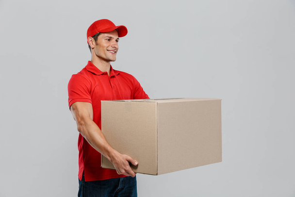 Young delivery man in hat smiling while posing with cardboard box isolated over white wall - Foto, Imagem