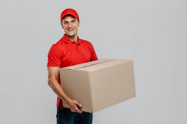 Young delivery man in hat smiling while posing with cardboard box isolated over white wall - Фото, зображення