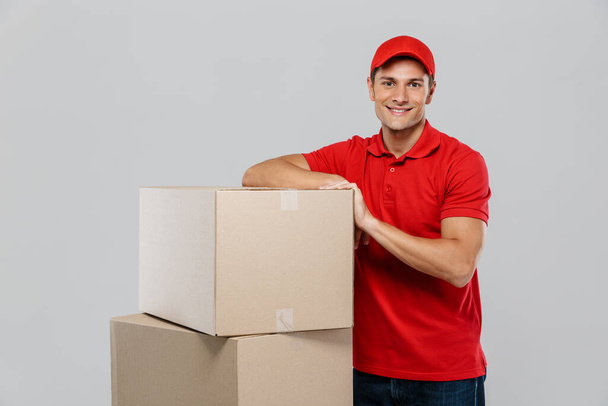 Young delivery man in hat smiling while posing with cardboard boxes isolated over white wall - Zdjęcie, obraz
