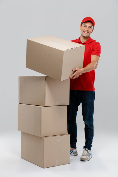 Young delivery man in hat smiling while posing with cardboard boxes isolated over white wall - Photo, image