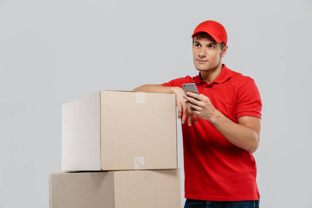 Young delivery man using cellphone while posing with cardboard boxes isolated over white wall - 写真・画像