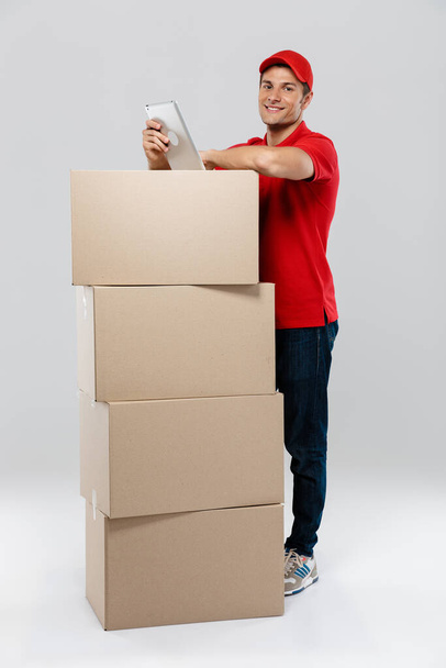 Young delivery man using tablet-computer while posing with cardboard boxes isolated over white wall - Foto, Imagem