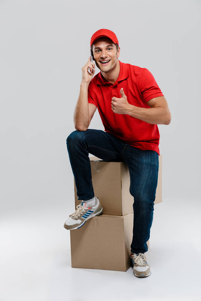 Young delivery man talking on cellphone while posing with cardboard boxes isolated over white wall - Fotoğraf, Görsel